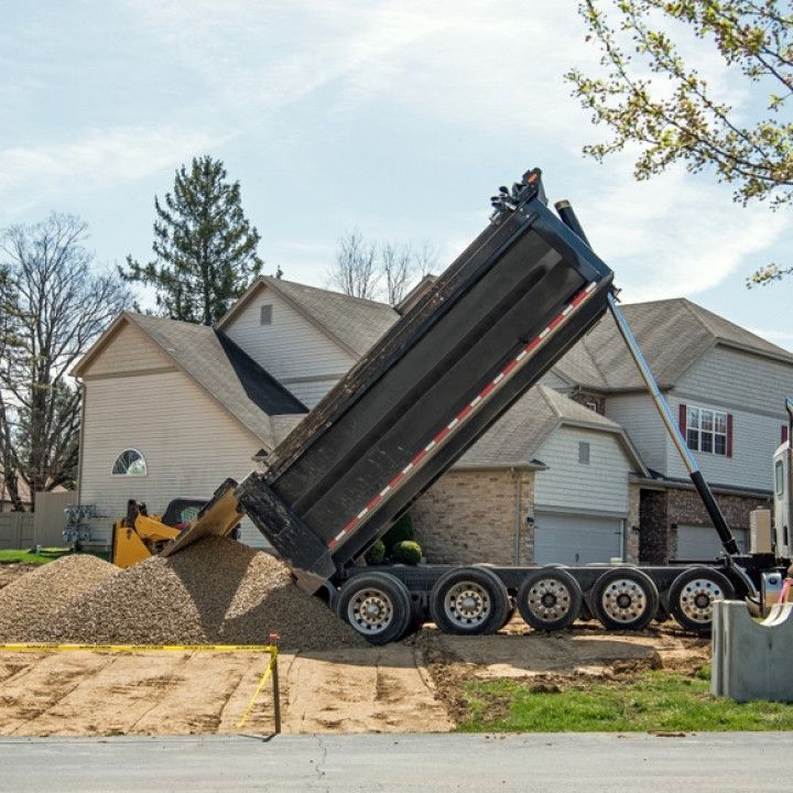 Dump truck unloading gravel in a suburban driveway beside a house under construction