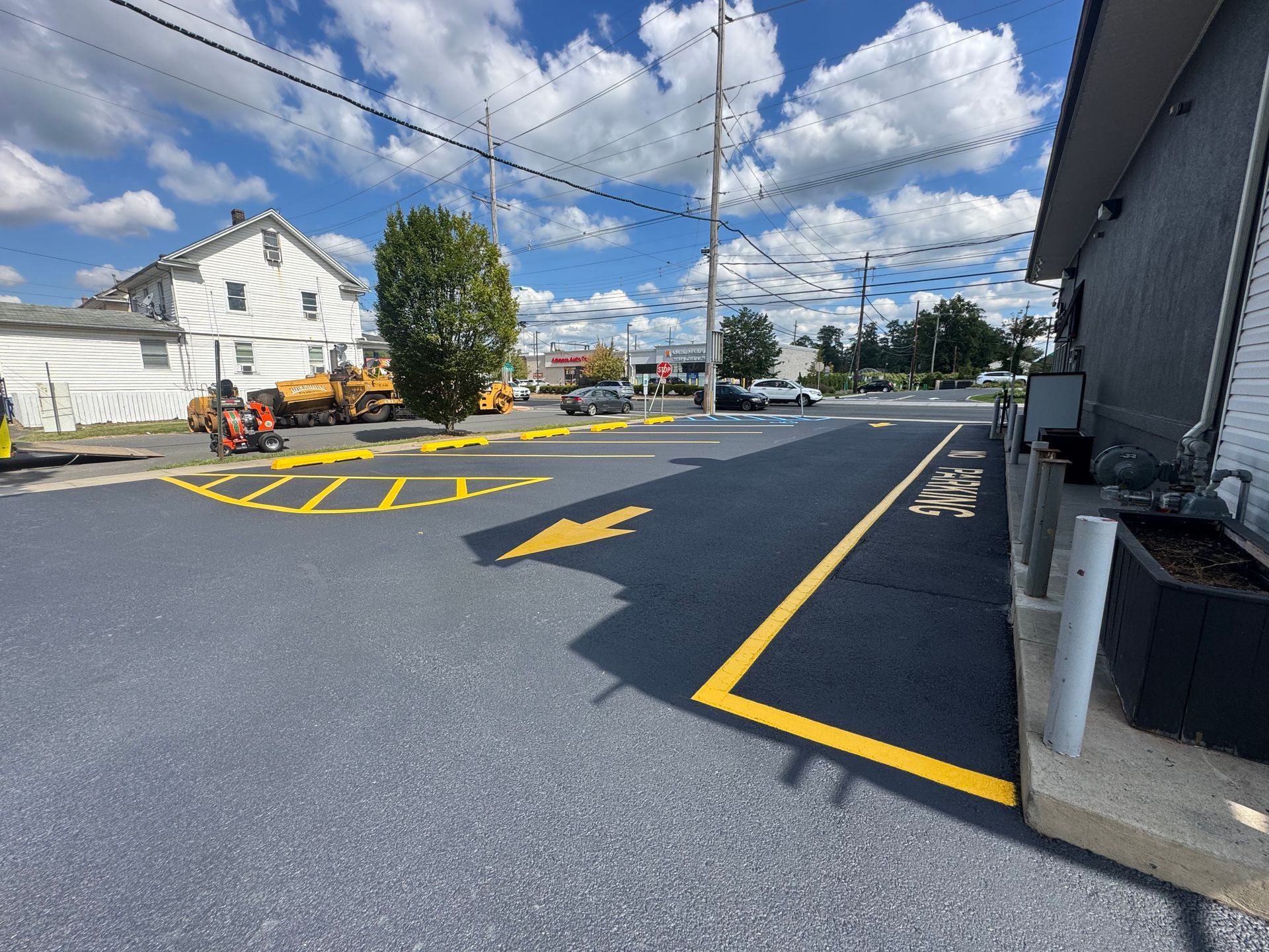Newly paved parking lot with yellow directional arrows and markings. Blue sky and commercial buildings in background.