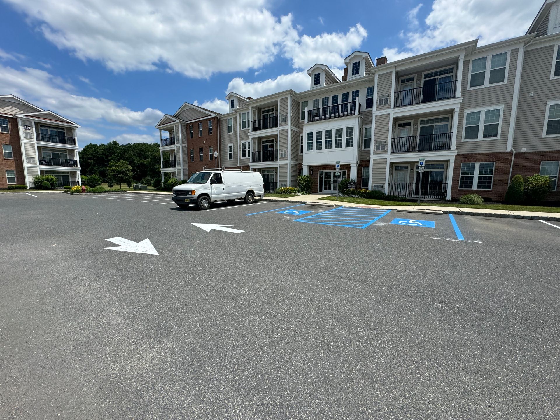 A white van parked in an asphalt parking lot with apartment buildings under a partly cloudy sky.