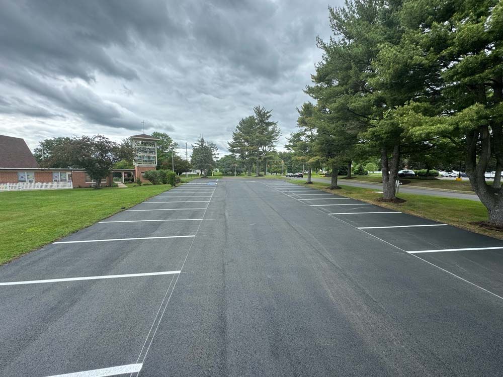 Newly paved parking lot with white painted parking space lines. Trees line the right side and a cloudy sky overhead.