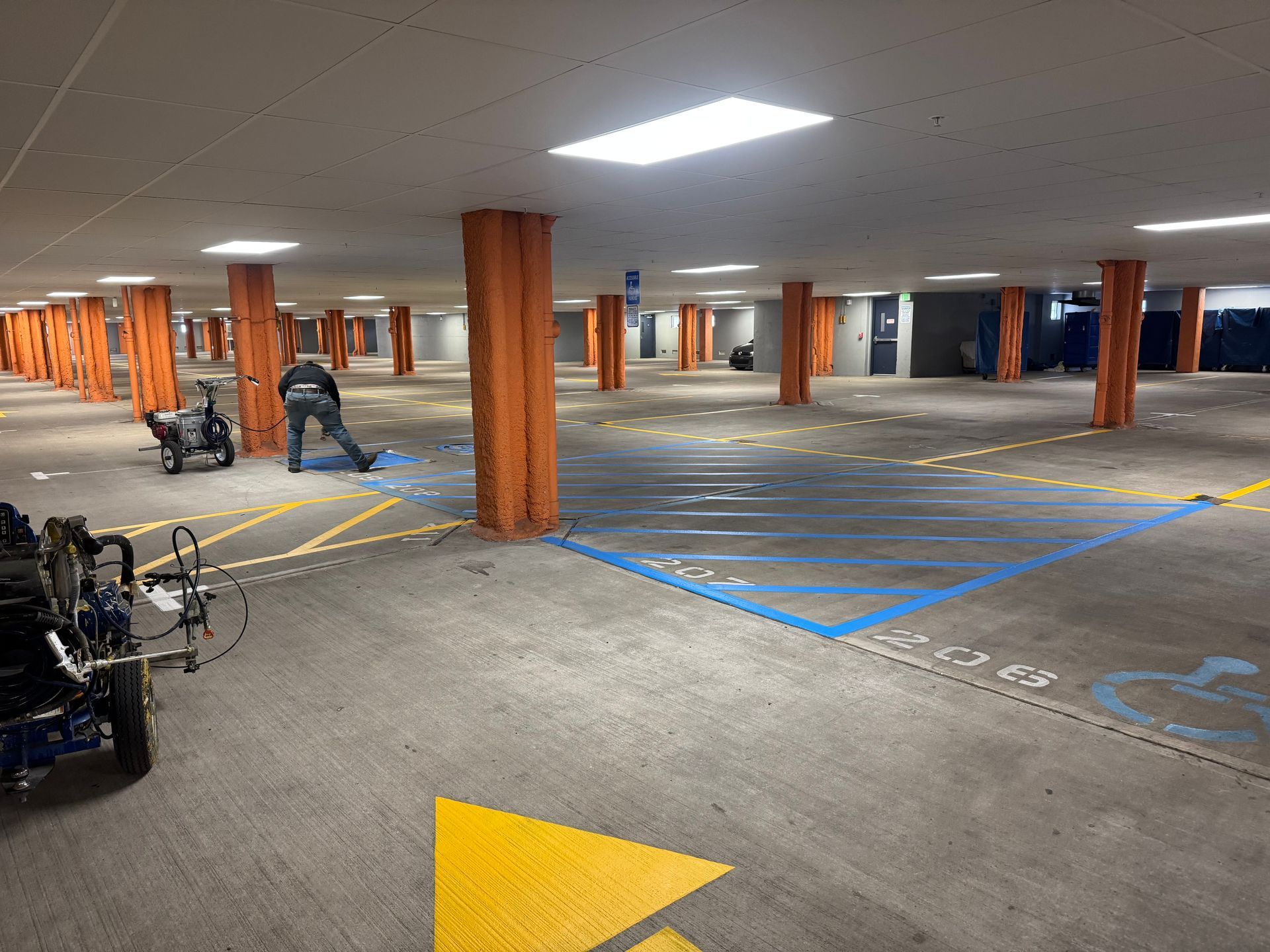 Parking garage with orange columns. A person paints parking space lines: blue, yellow, and white.
