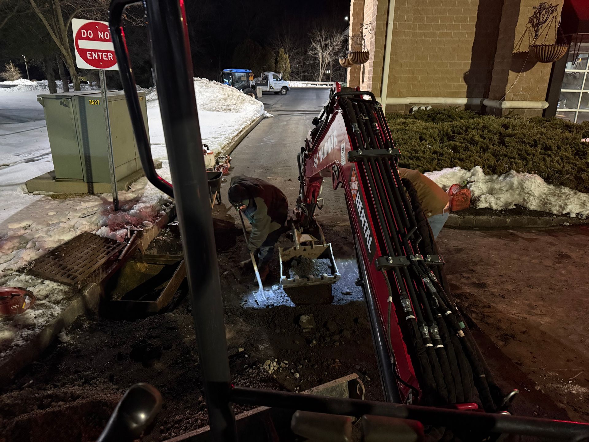 Snowy roadside construction scene with a red utility trenching machine beside a building at night
