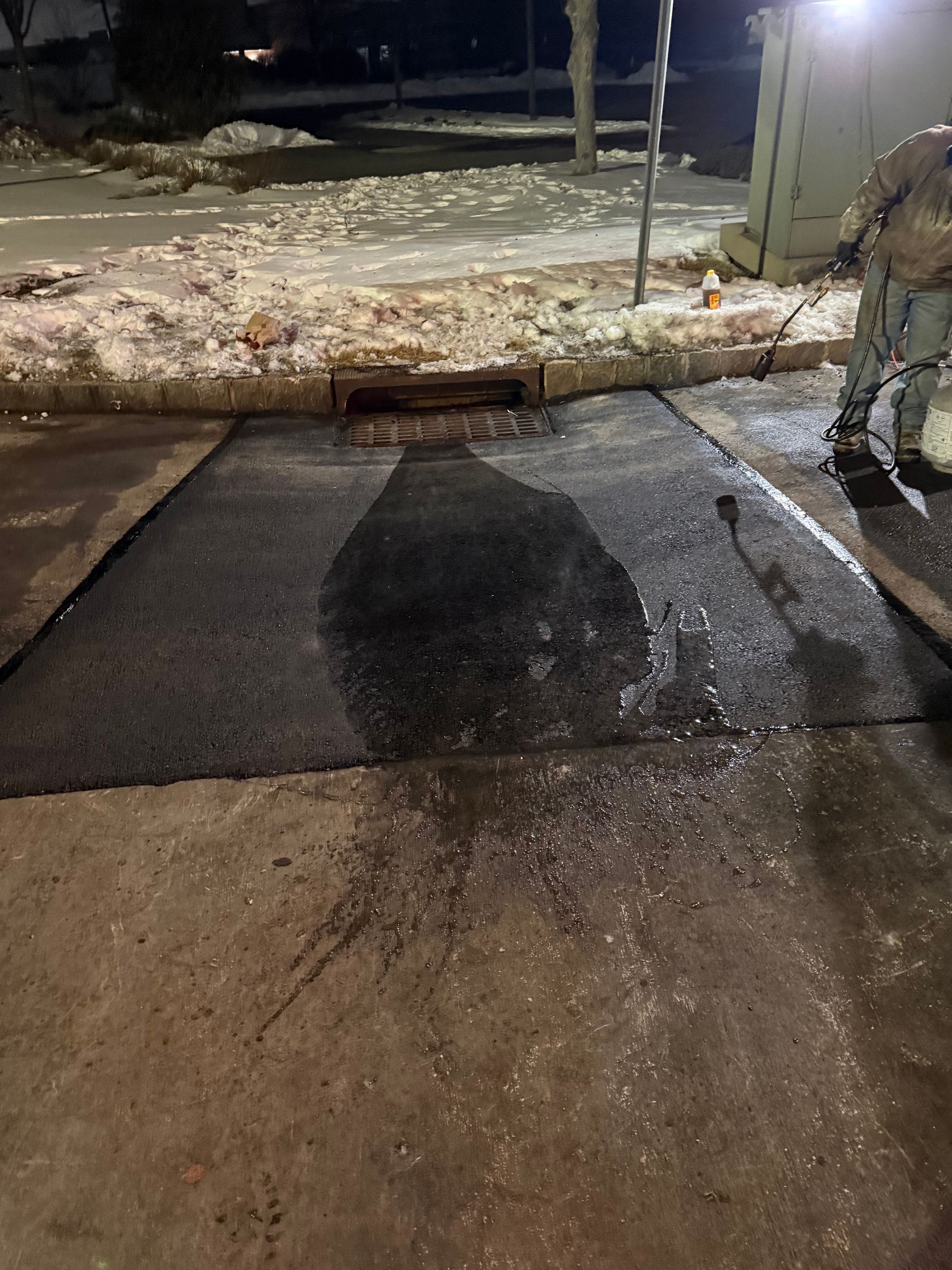 Snow-covered curb blocking a wet driveway at night, with a person standing at the right edge