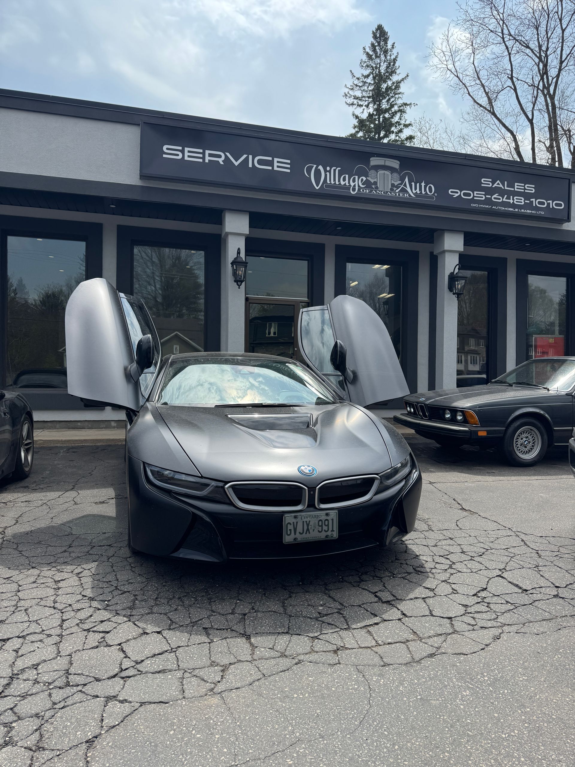 Black BMW i8 with scissor doors open outside a service shop, with a black car parked nearby.