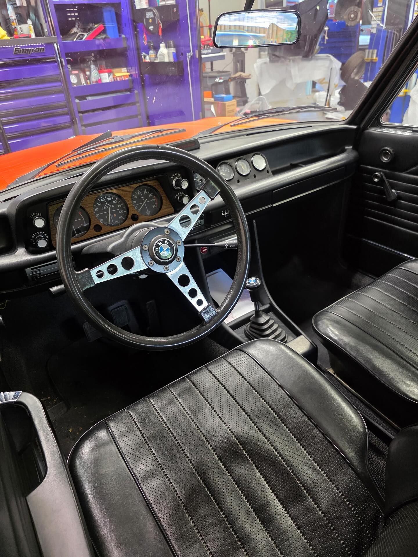 Interior of a classic muscle car with black seats, orange dash, and custom steering wheel in a garage