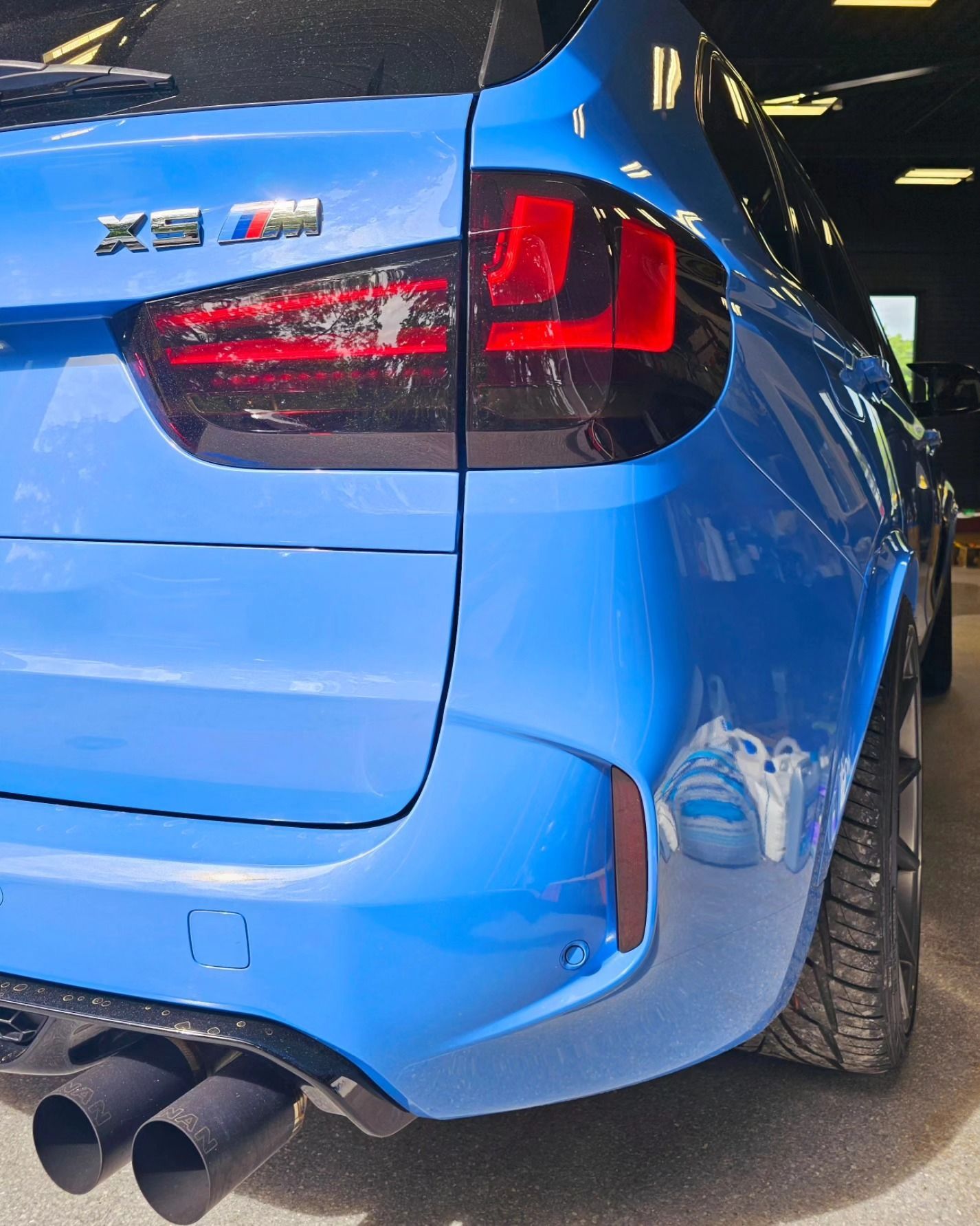 Rear-left of a blue BMW X5 M with red taillight and twin exhausts inside a garage.