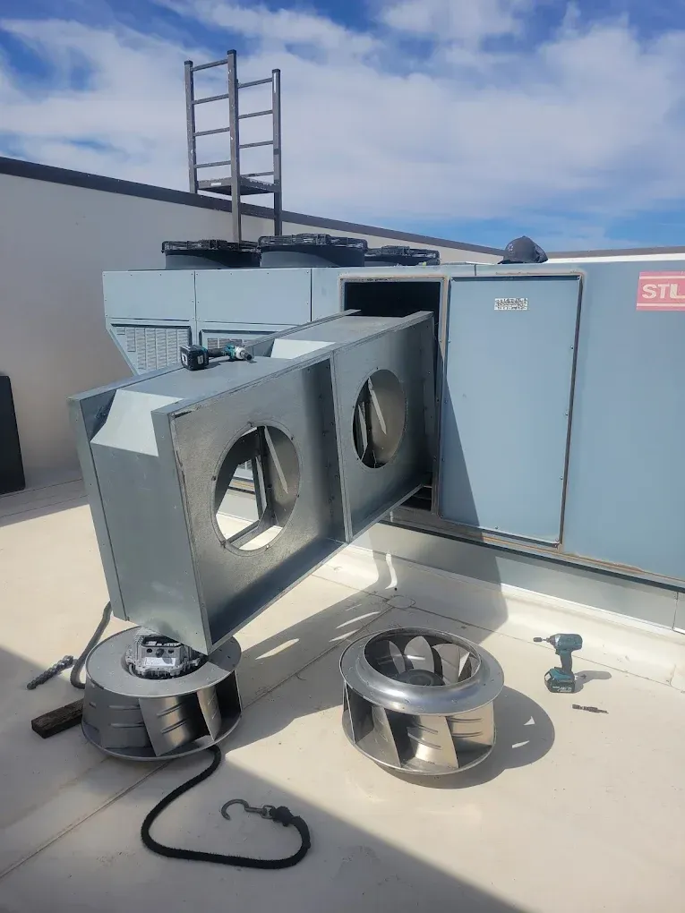 Metal HVAC equipment on a rooftop, with ducts, fans, and a ladder against a blue utility unit.