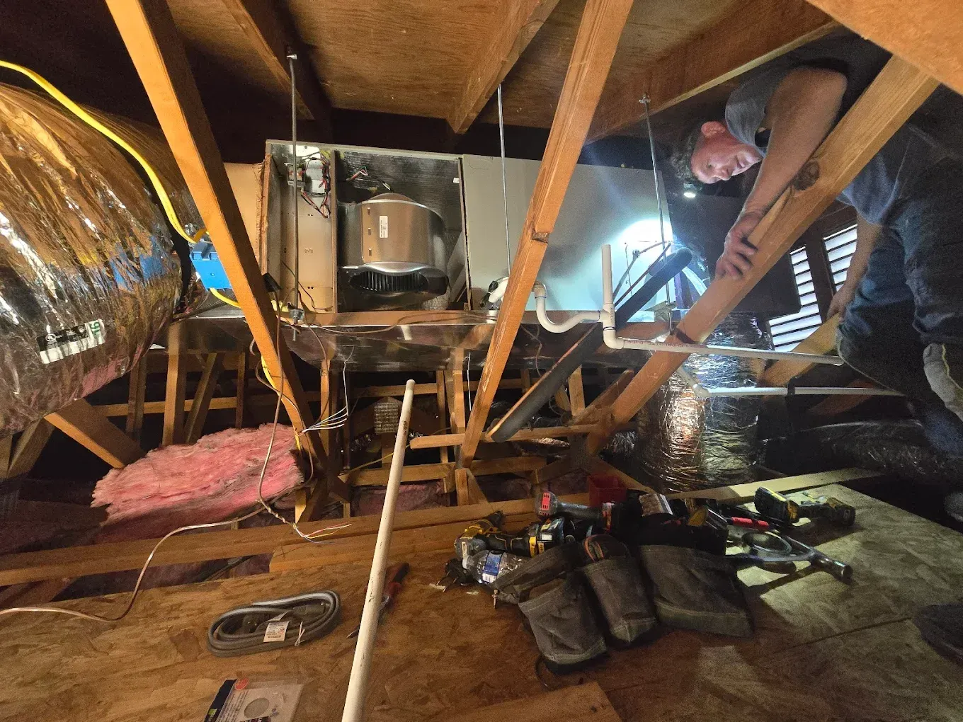 Attic crawlspace with exposed insulation, wooden beams, tools, and a person inspecting the roof on the right.