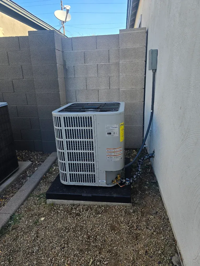 Outdoor air conditioner unit beside a stucco wall and cinderblock fence, on gravel with a black base.