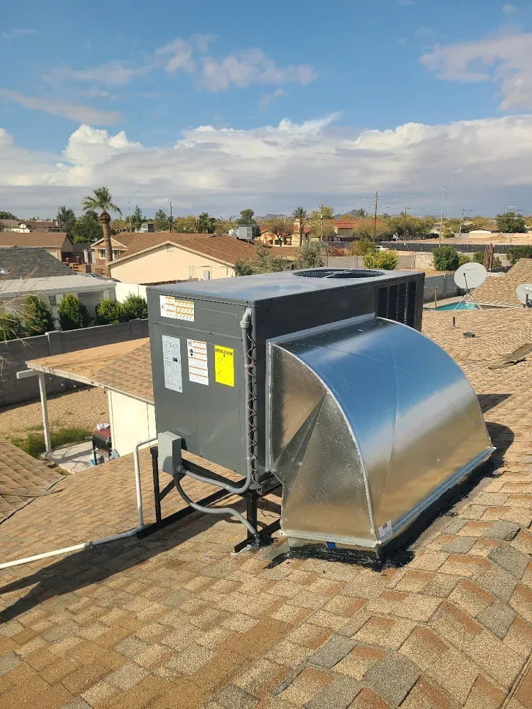 Rooftop HVAC unit with metal ducting on a shingled roof under a blue sky.