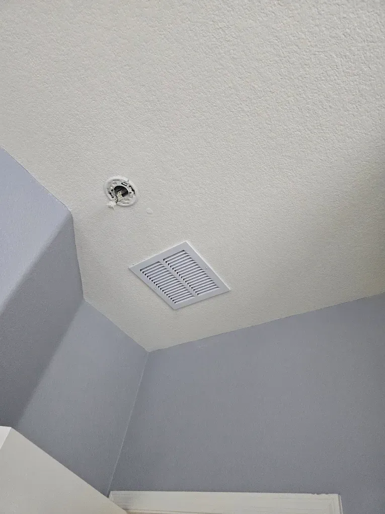 Ceiling corner with a white vent and recessed light on light gray walls.