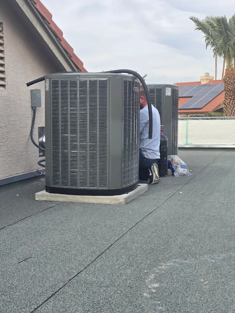 Worker kneeling beside rooftop HVAC units on a flat roof near a house with solar panels.