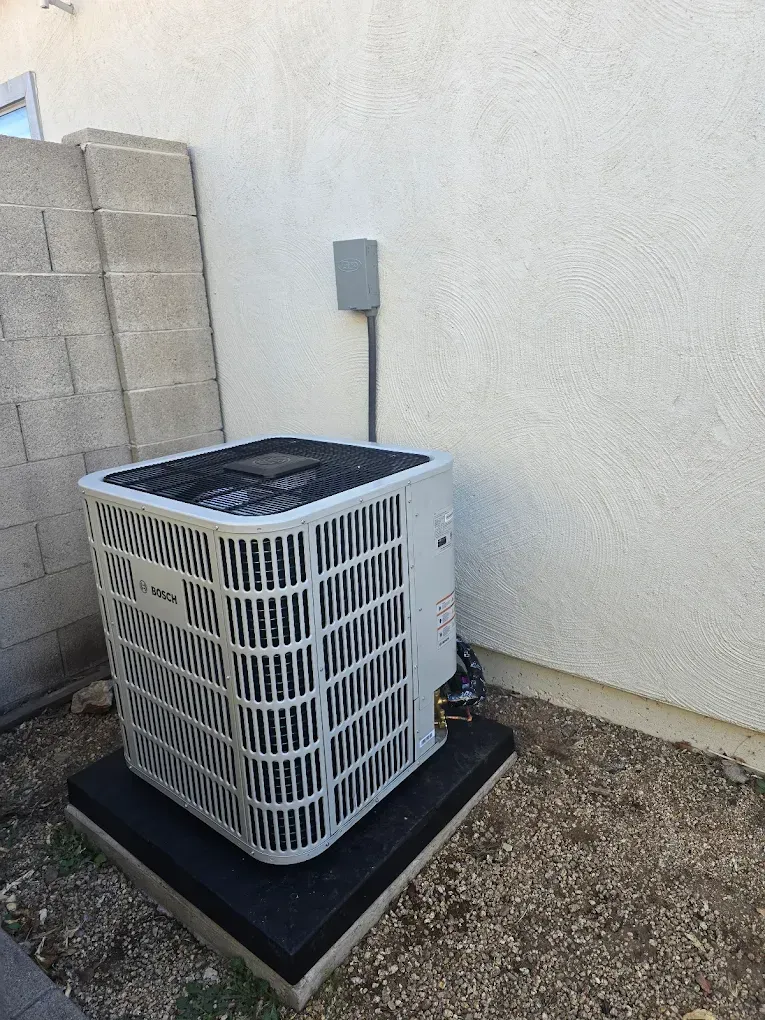 Outdoor air conditioner unit on a black pad beside a beige wall and cinder block fence.