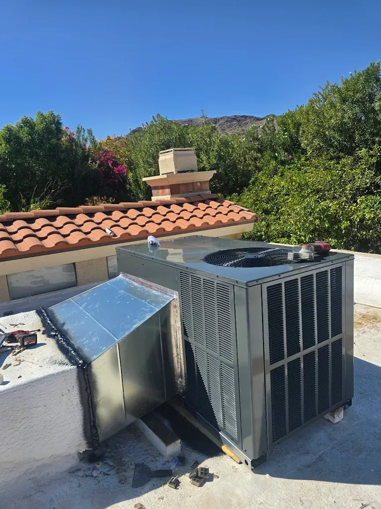 Rooftop HVAC unit and ductwork beside a red-tile roof under a clear blue sky