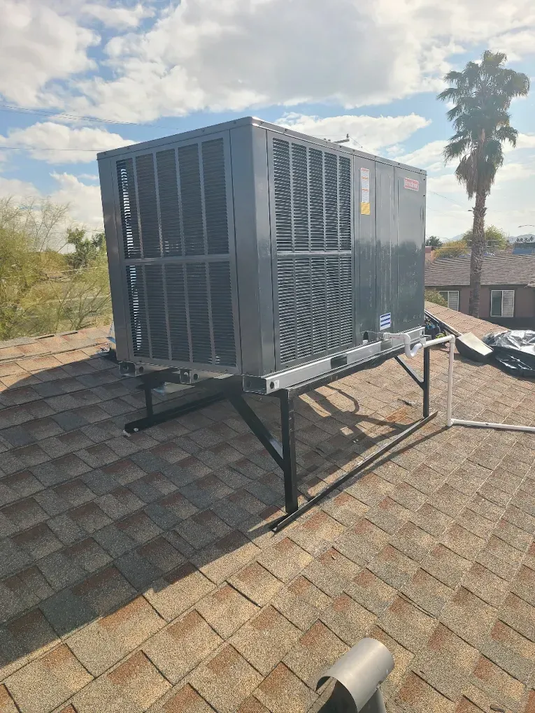 Gray rooftop HVAC unit on black metal stand against a sunny sky and palm tree.
