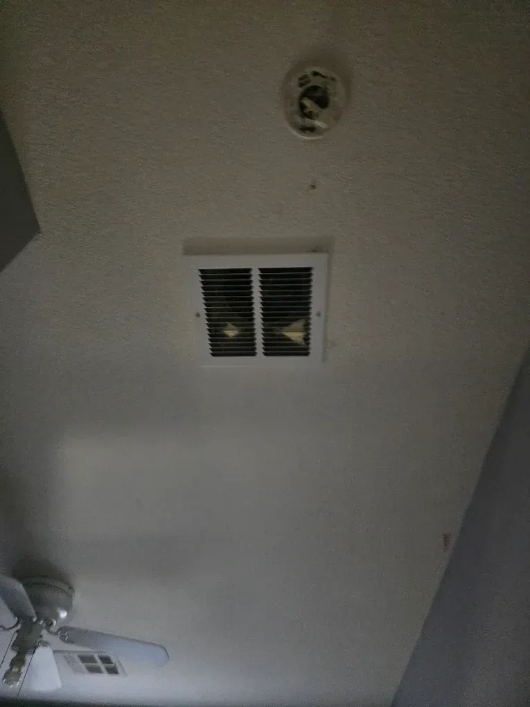 Ceiling with a small vent, smoke detector, and part of a ceiling fan in a room