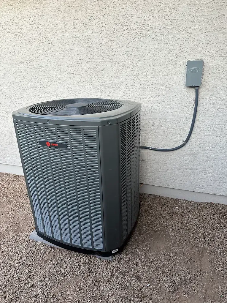 Outdoor air conditioner unit beside a beige stucco wall with a connected power box and gravel ground