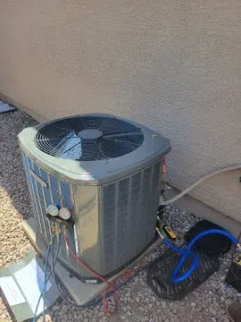 Outdoor air conditioner unit beside a wall with connected hoses and a blue bucket on a gravel pad