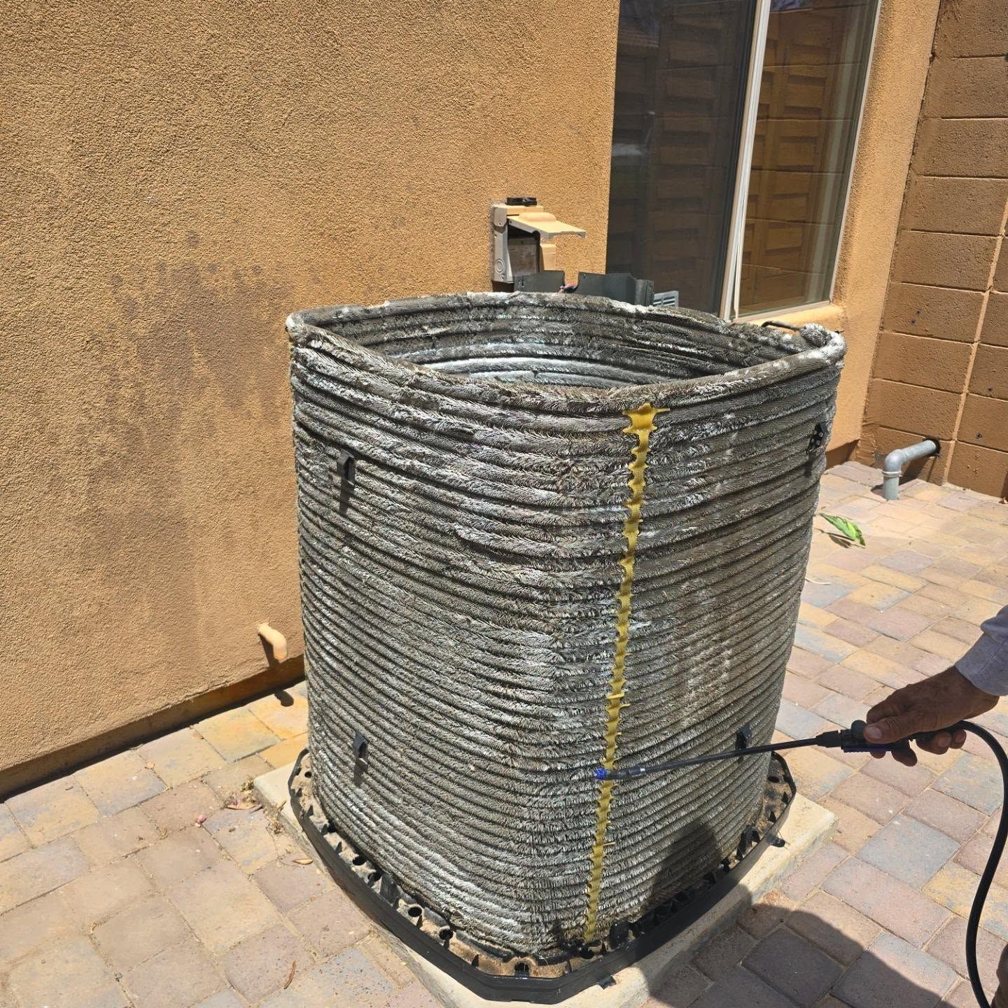 Large gray woven outdoor unit cover beside a tan wall, with a hose attached on a paved patio.