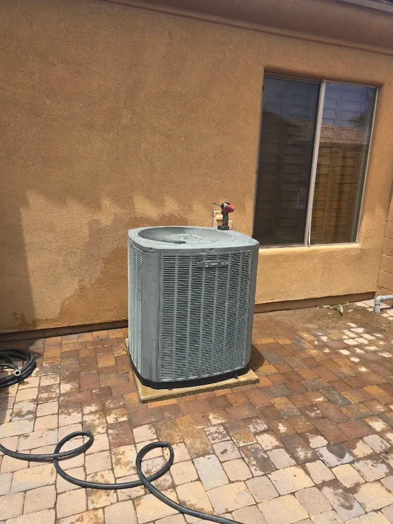 Outdoor air conditioner unit beside a tan stucco wall on a brick patio, with a hose on the ground.