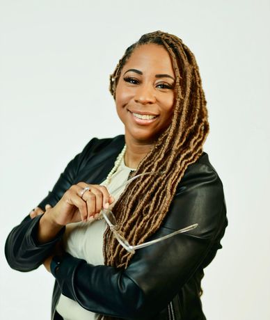 A woman with dreadlocks is wearing a black leather jacket and holding a pair of glasses.