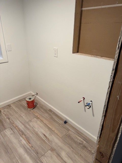 Small unfinished room with white walls, wood-look flooring, a paint bucket, and exposed plumbing fixtures