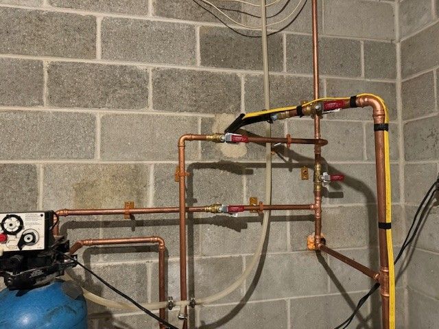 Copper pipes and valves mounted on a cinderblock wall beside a blue tank in a utility room.