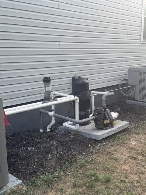Pool equipment on the side of a house that includes a pump, filter, and plumbing on a concrete pad