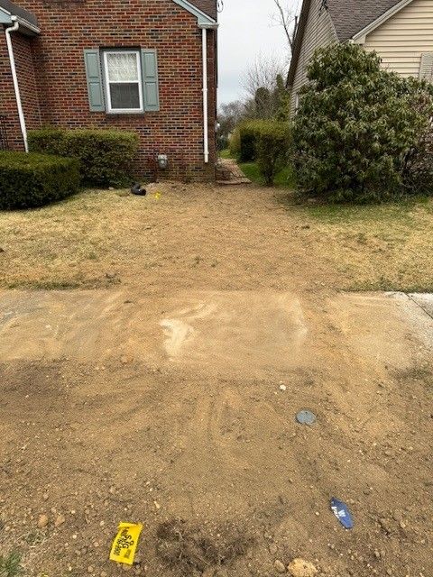 Front yard with a bare dirt path leading to a brick house, bordered by grass and shrubs.