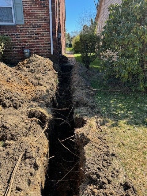 Narrow trench dug beside a brick house, with piles of excavated dirt along the edge.