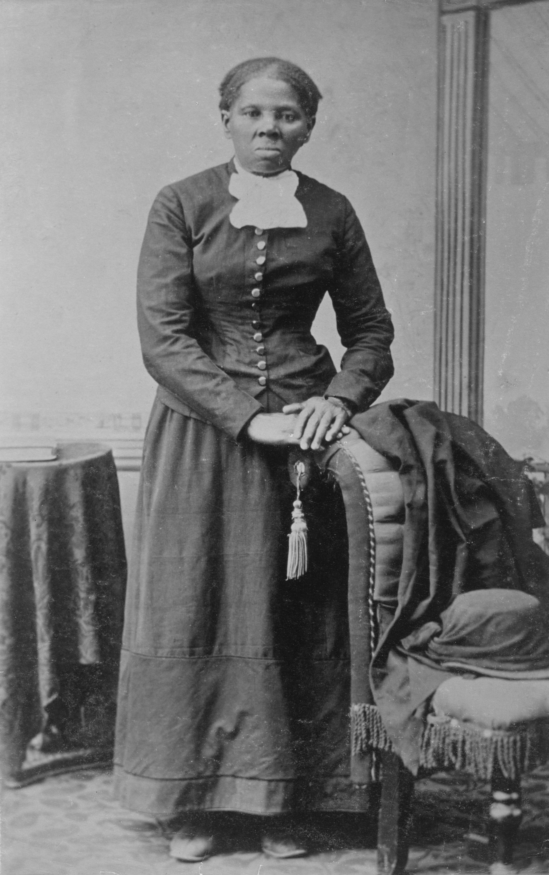 Full-length portrait of a person standing indoors beside a draped chair and small round table