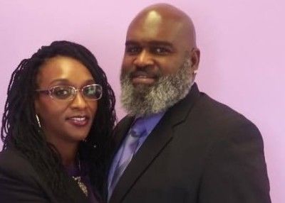 Two people posing together against a purple wall, wearing formal attire and smiling.