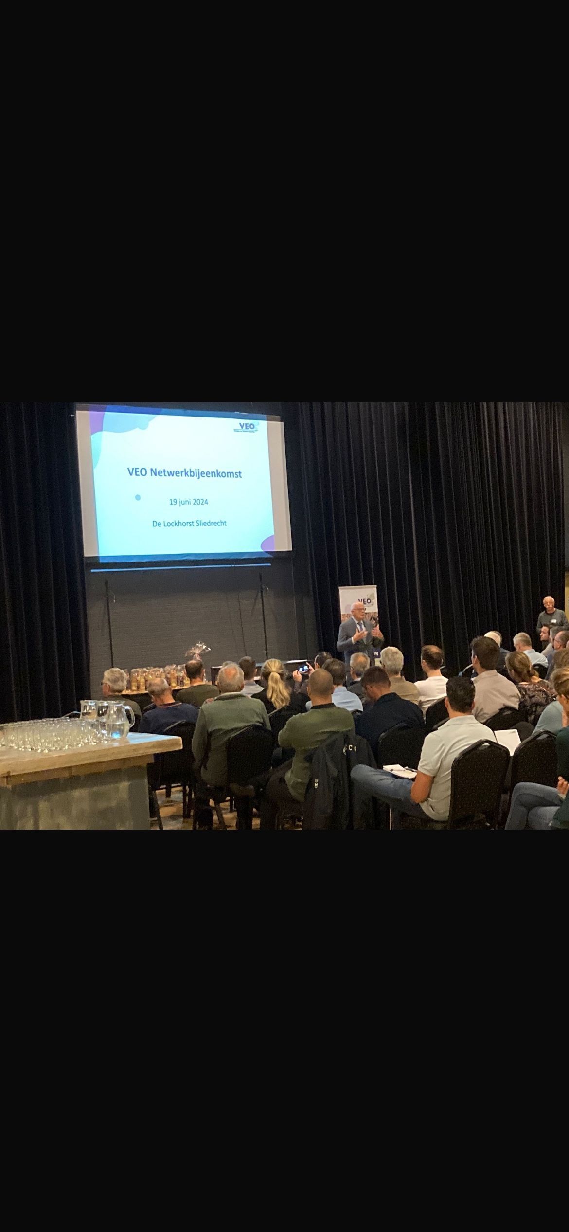 Een groep mensen zit tijdens een conferentie voor een groot scherm.