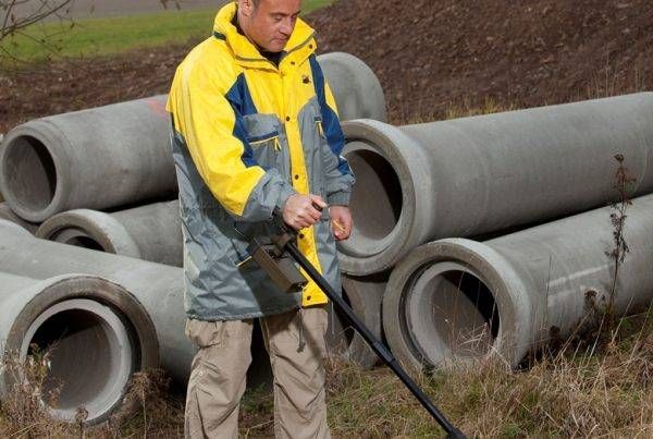 Een man in een geel-blauw jasje staat voor een stapel betonnen buizen.