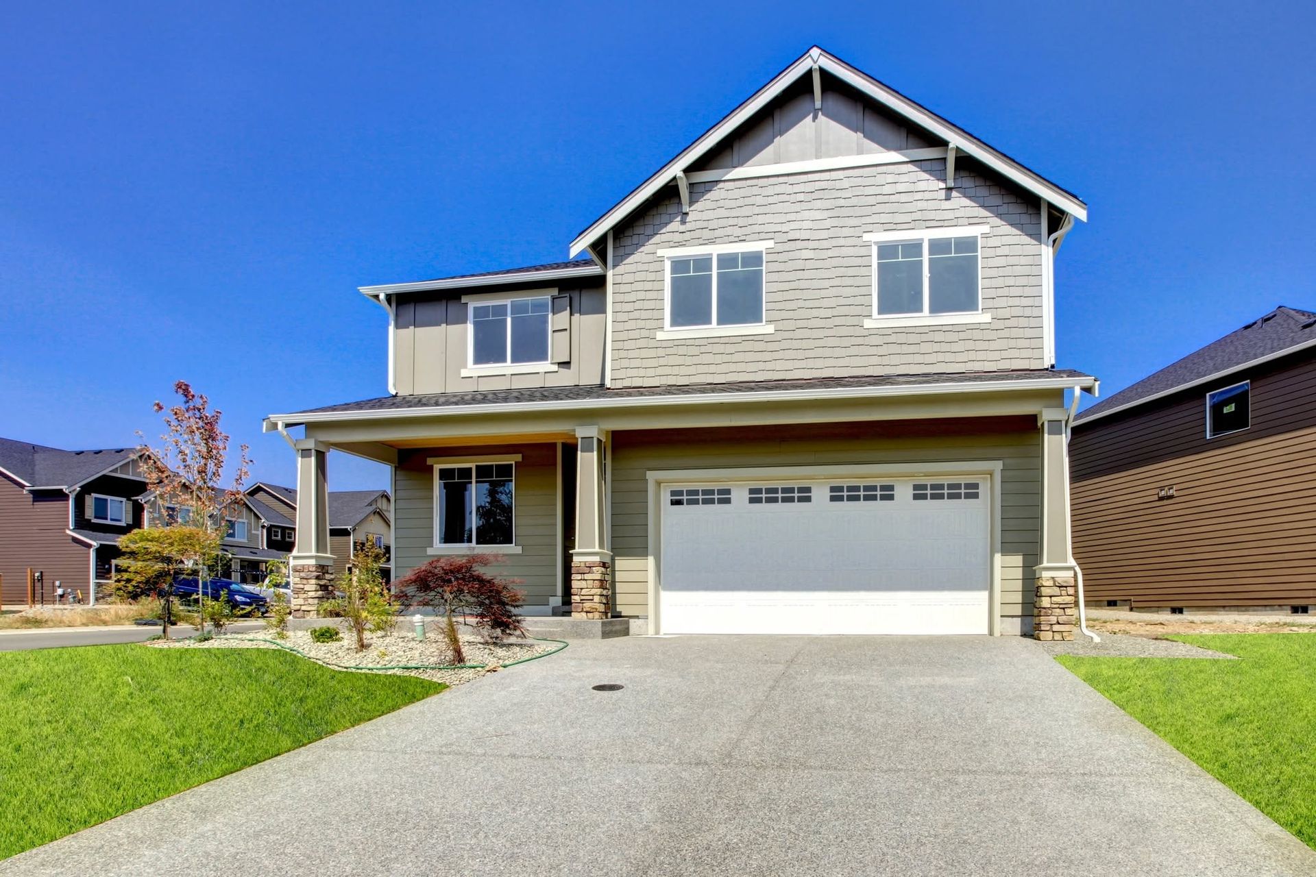 Two-story beige house with a front porch, garage, and a green lawn under a bright blue sky