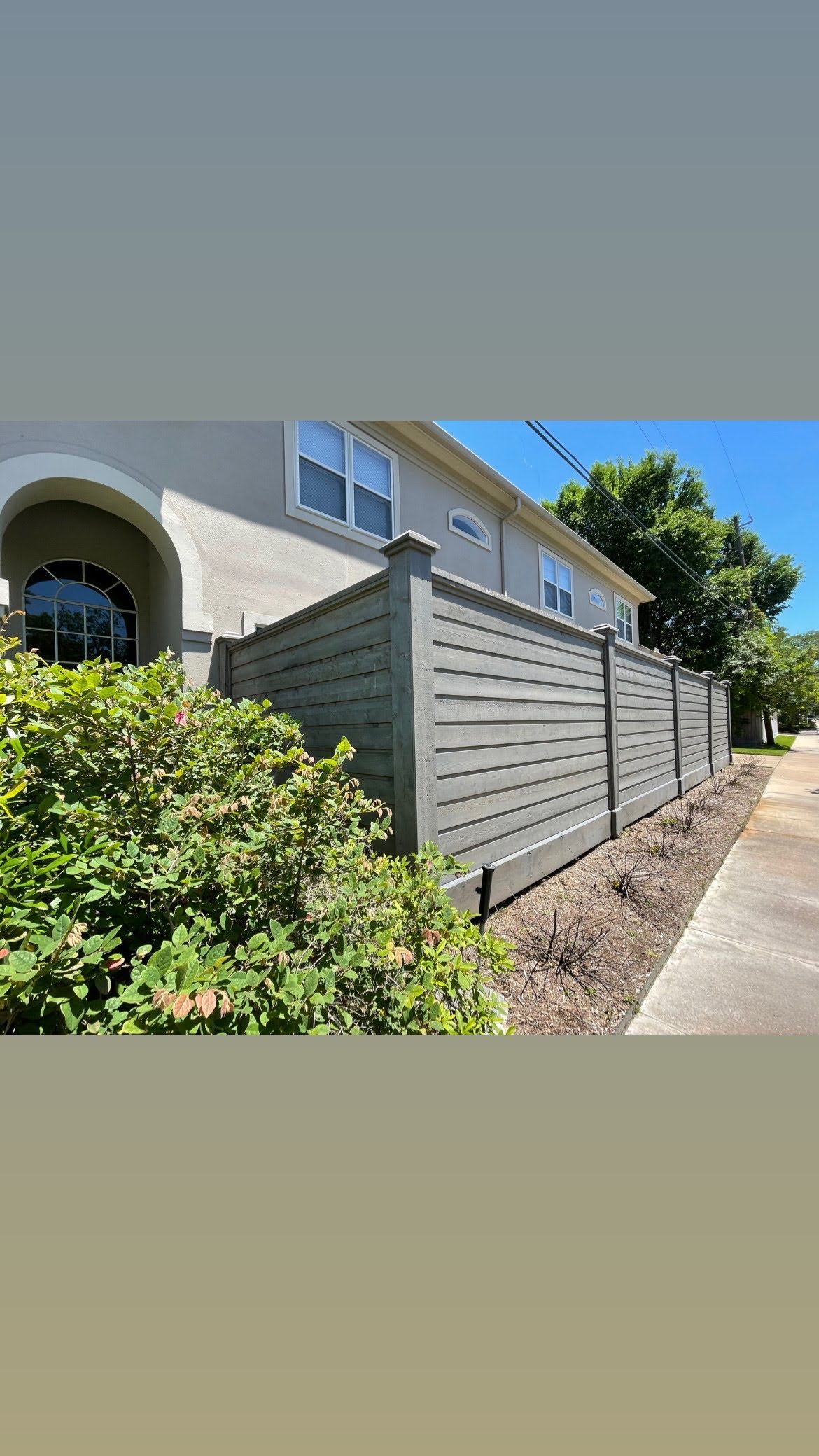 Gray horizontal slat fence in front of a light-colored building with green foliage and sidewalk.