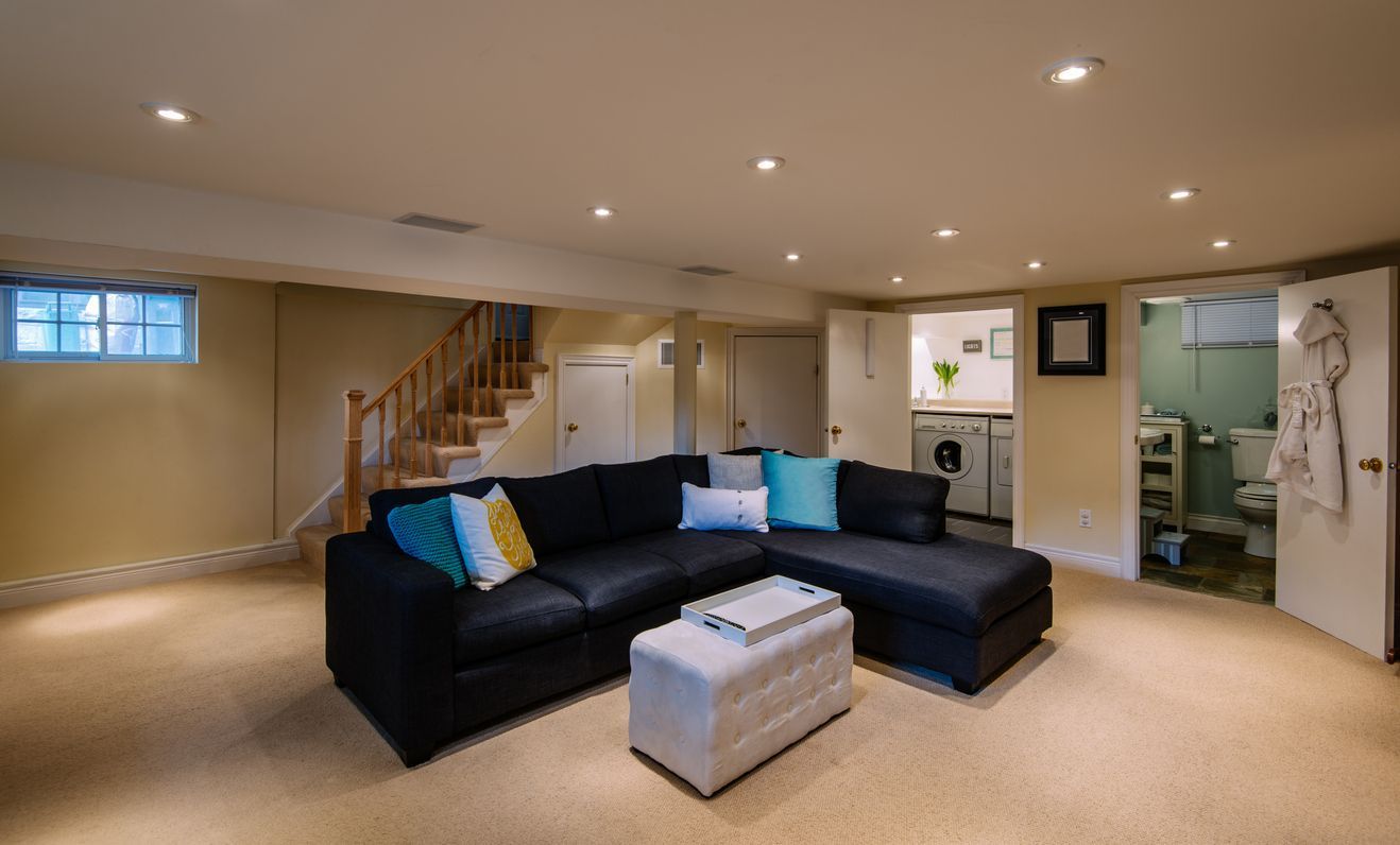 A furnished basement featuring a black sectional sofa, a beige ottoman, a staircase, and open doorways to a utility area.
