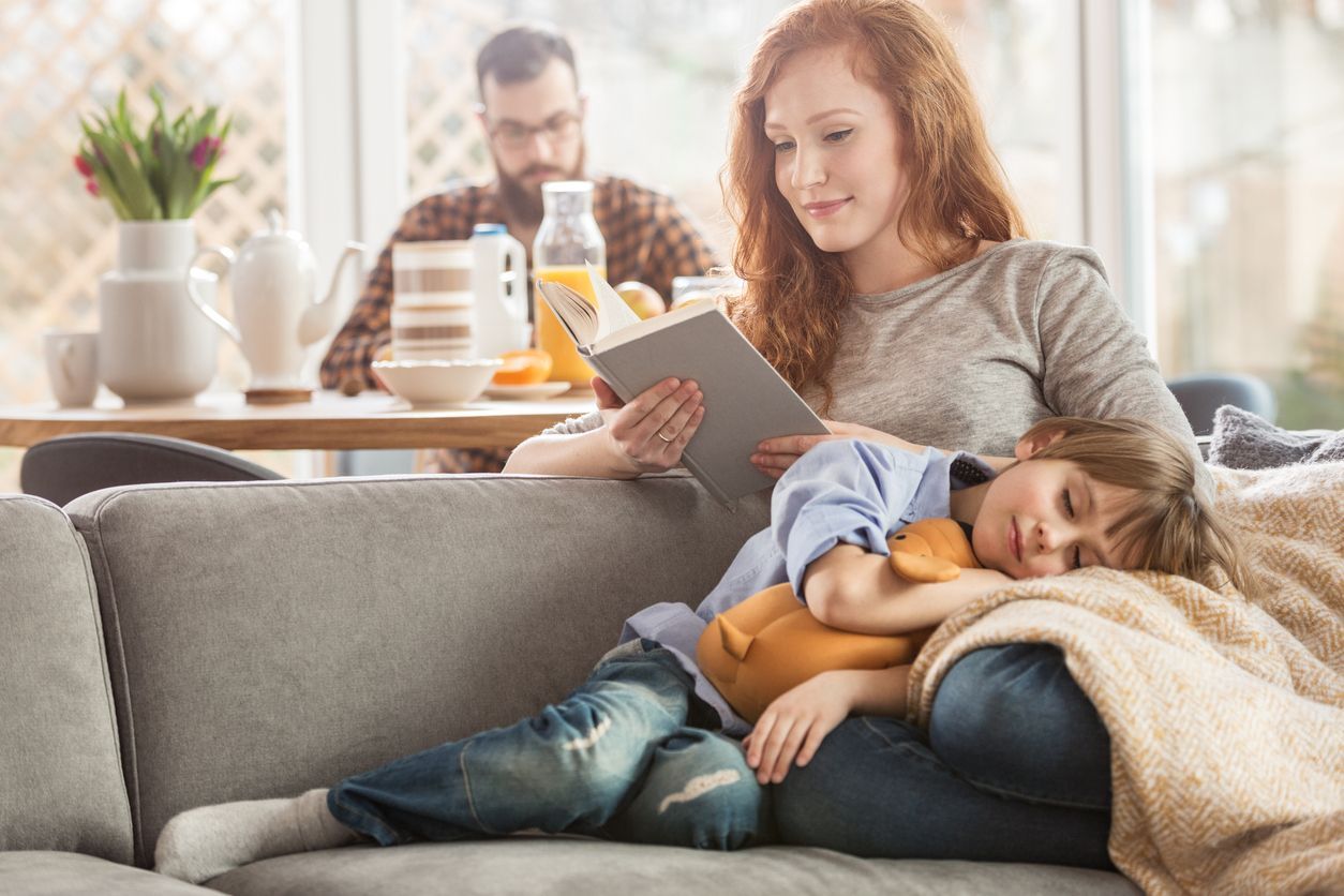 A person reads a book while a child sleeps on their lap on a sofa, with another person sitting at a table in the background.