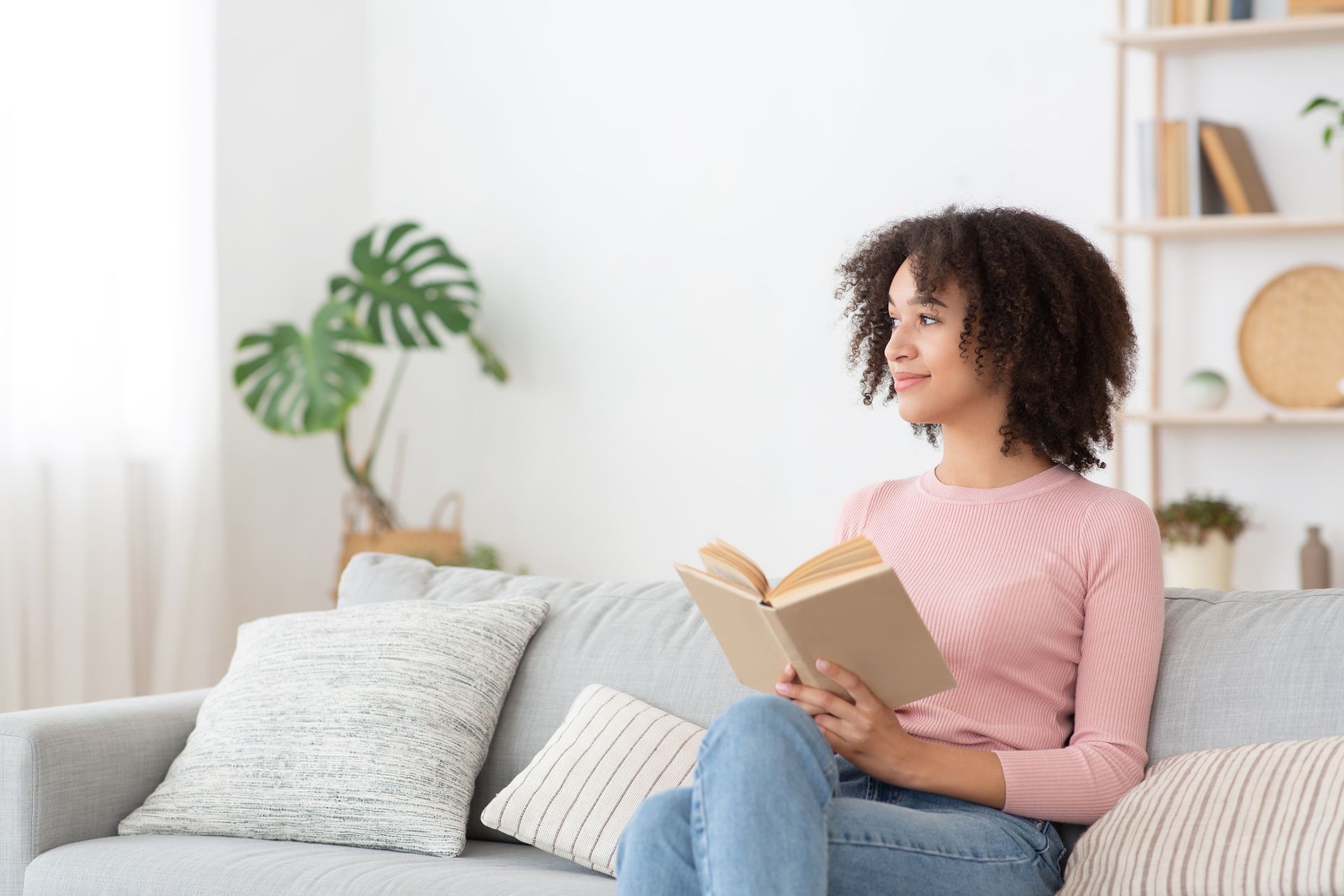 A person with curly hair wearing a pink long-sleeved shirt sits on a grey couch holding an open book and looking away.