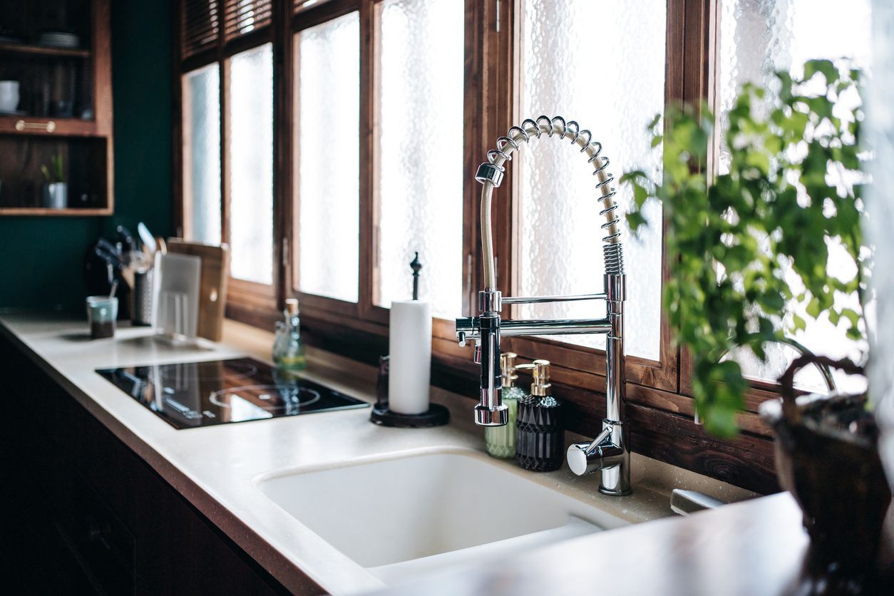 A white kitchen sink with a chrome gooseneck faucet, paper towel roll, and small potted plant set against window panes.