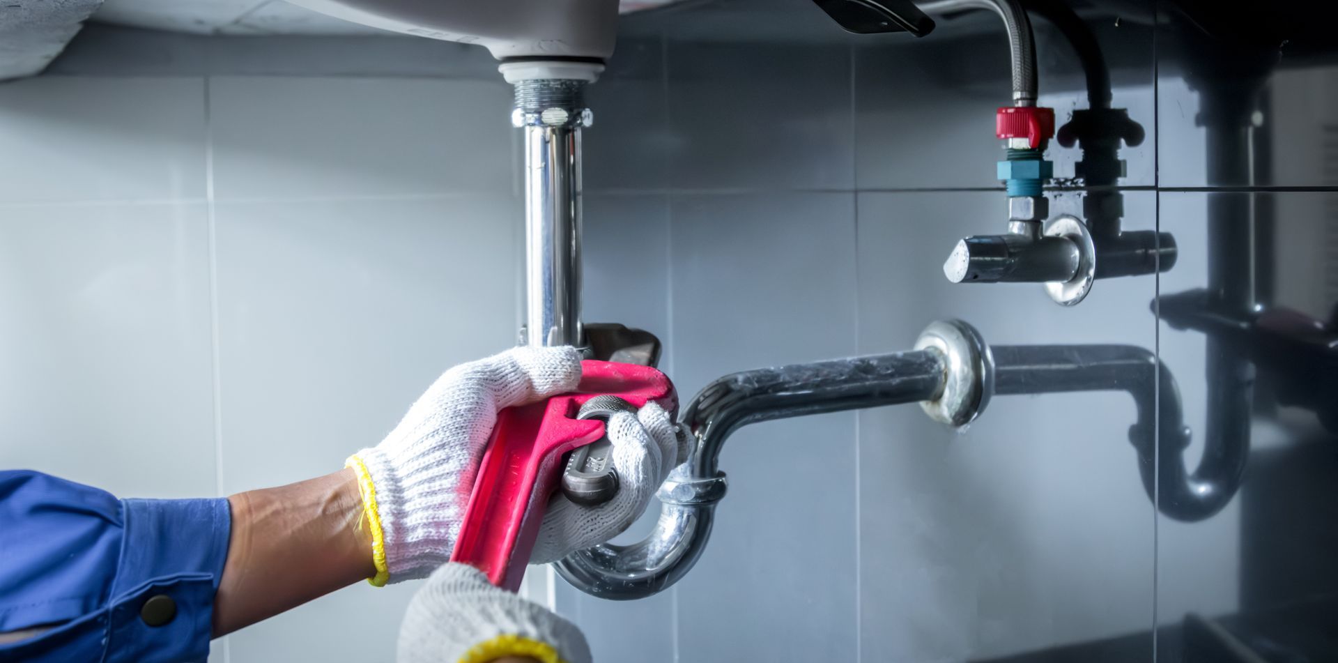 A person wearing white gloves uses a pink wrench to tighten a metal pipe under a sink.