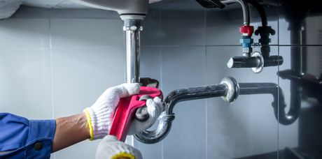 A person wearing gloves uses a red pipe wrench to adjust the metal P-trap piping under a bathroom or kitchen sink.