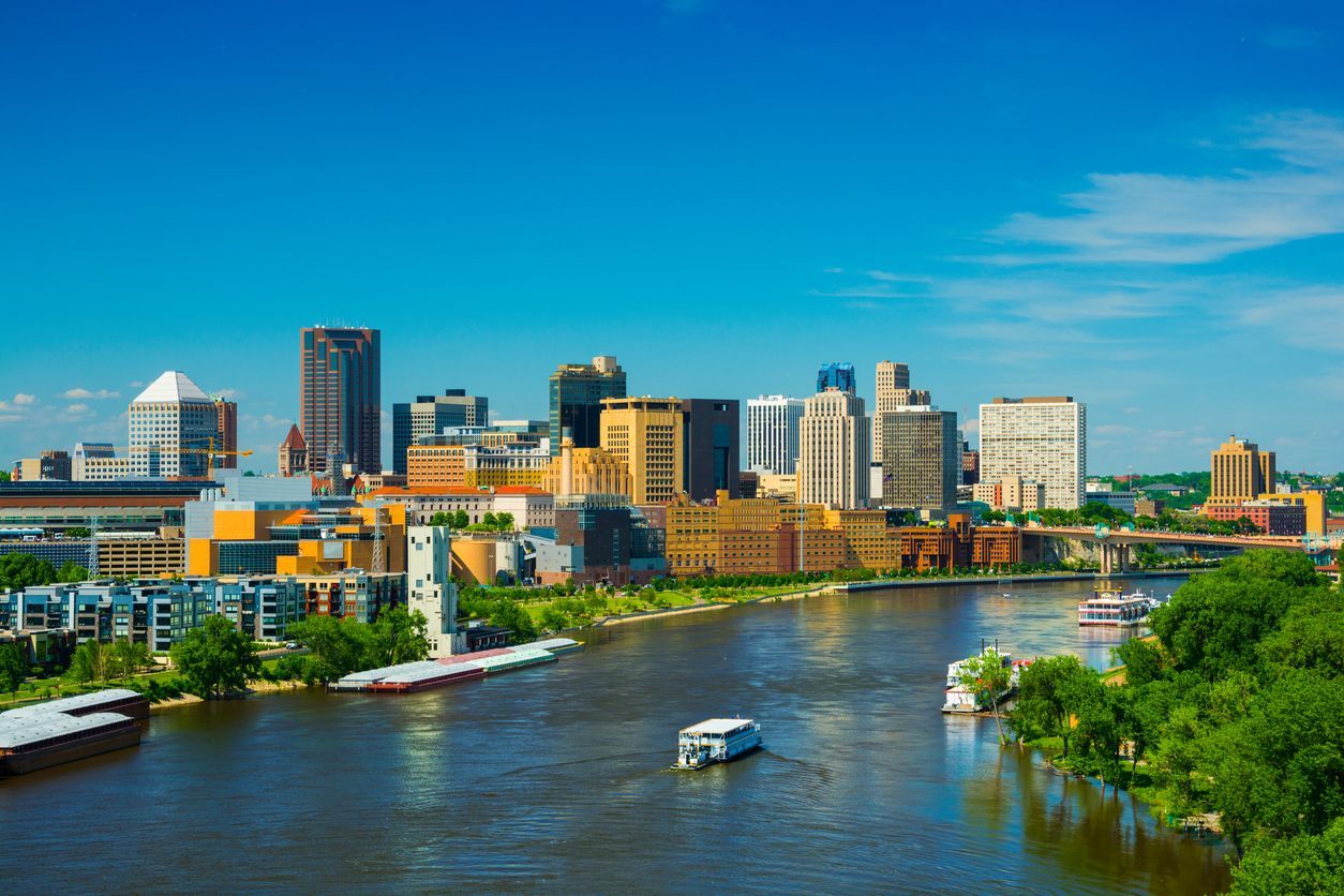 A vibrant city skyline on a river with a boat traveling through the center under a clear blue sky.
