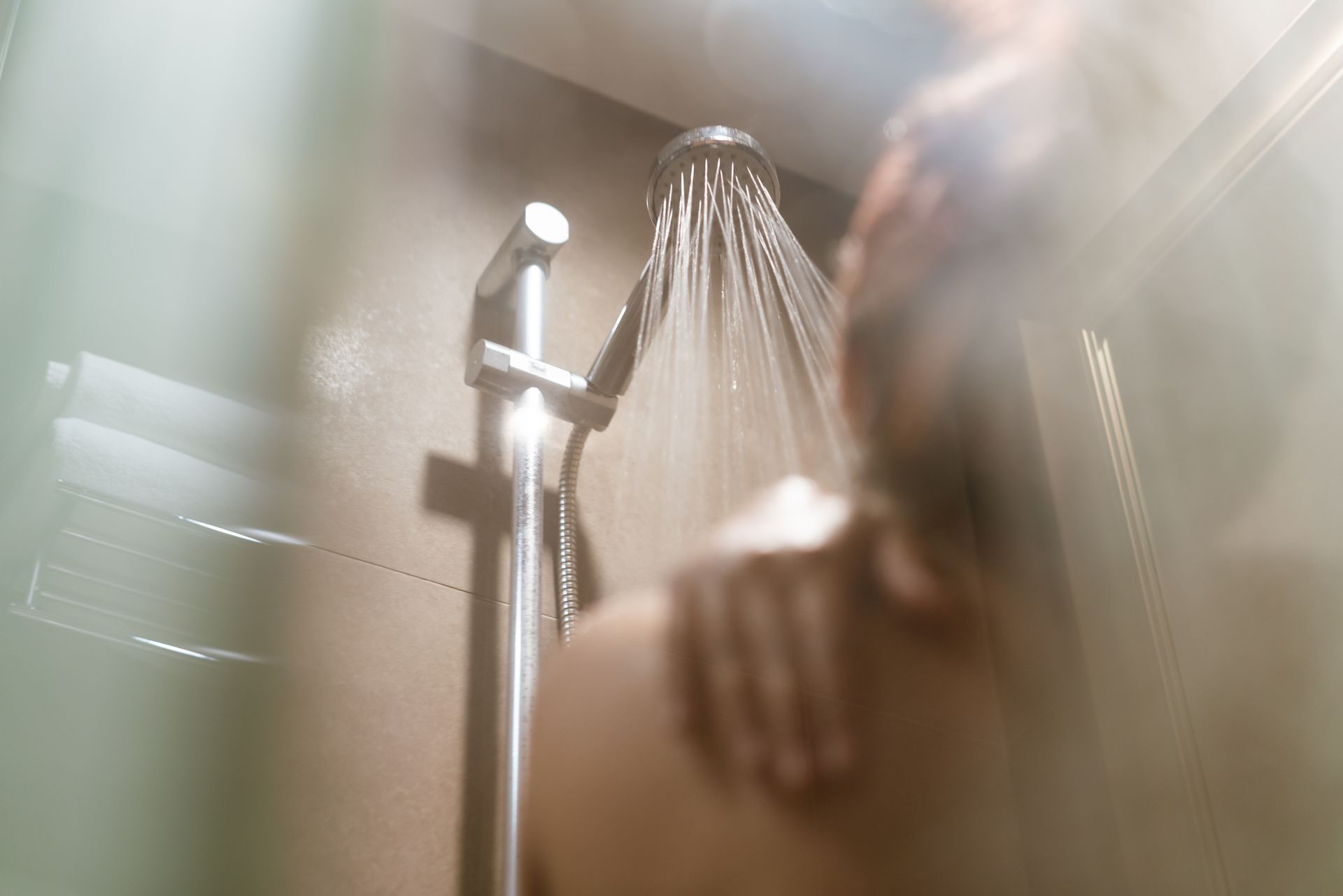 A person’s shoulder and hand visible behind a glass shower door as water cascades from a modern showerhead.