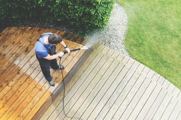 Person pressure washing a wooden deck beside a green lawn and hedge