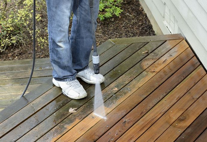 Person pressure-washing a wooden deck near a house wall.