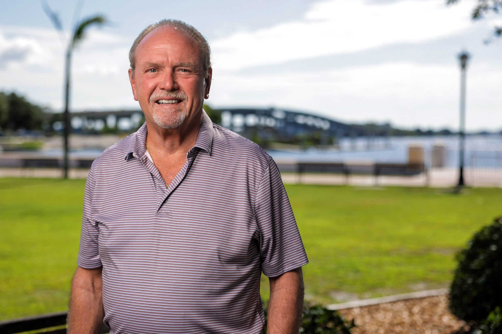 Man standing outdoors near a waterfront, wearing a striped shirt and smiling at the camera.