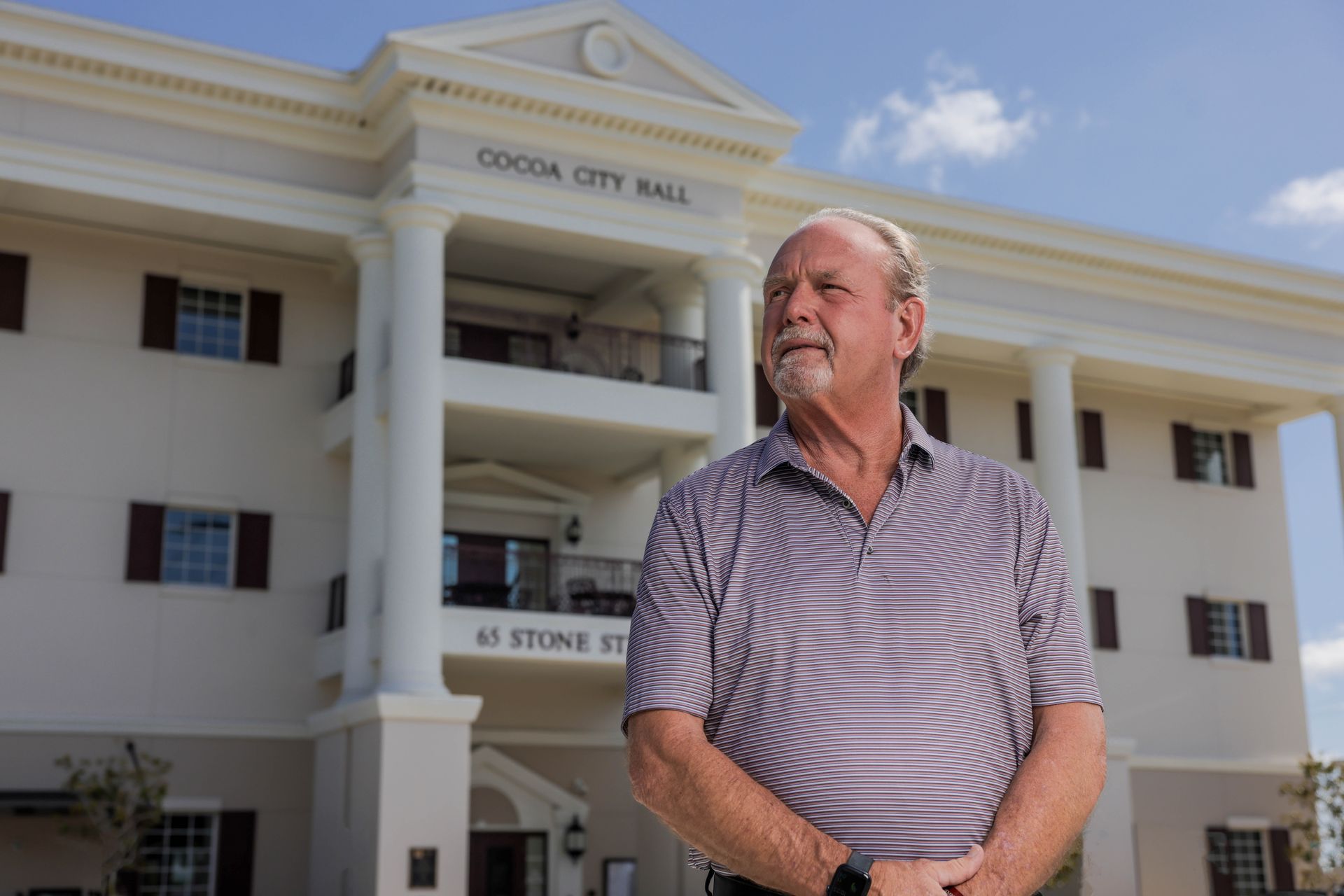 Man standing in front of a white city hall building with columns and a sign reading “City Hall.”