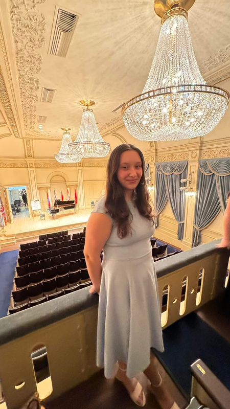 Woman in a light gray dress standing on a balcony overlooking a chandelier-lit theater interior.