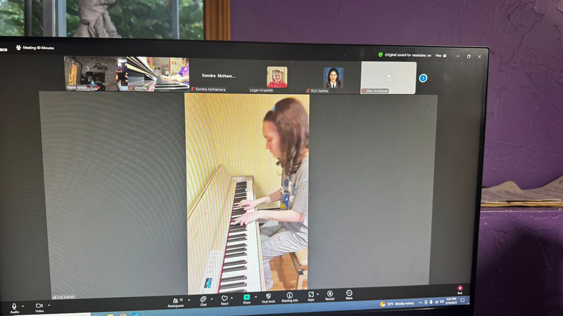 Laptop screen showing a video call with a woman playing a piano in a room.
